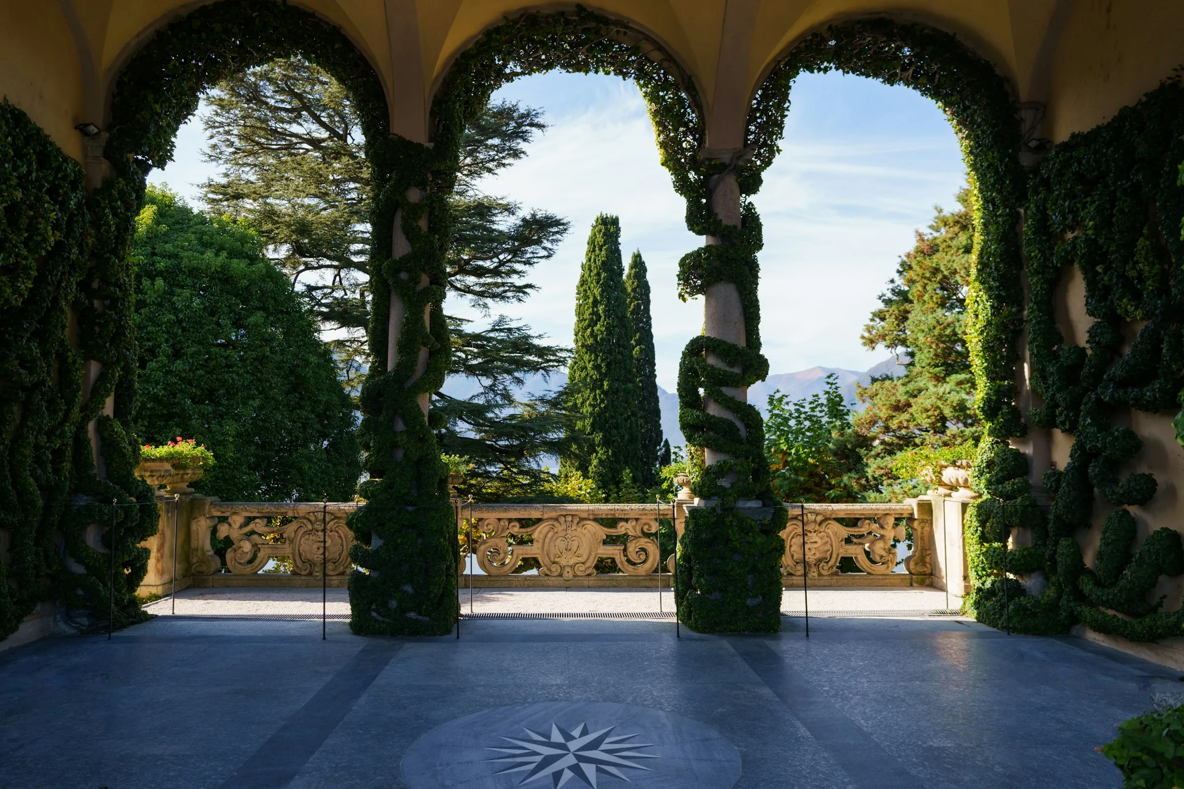 Loggia aux colonnes couvertes de lierre ouvrant sur une terrasse à balustrade de pierre, cyprès et cèdres au-delà